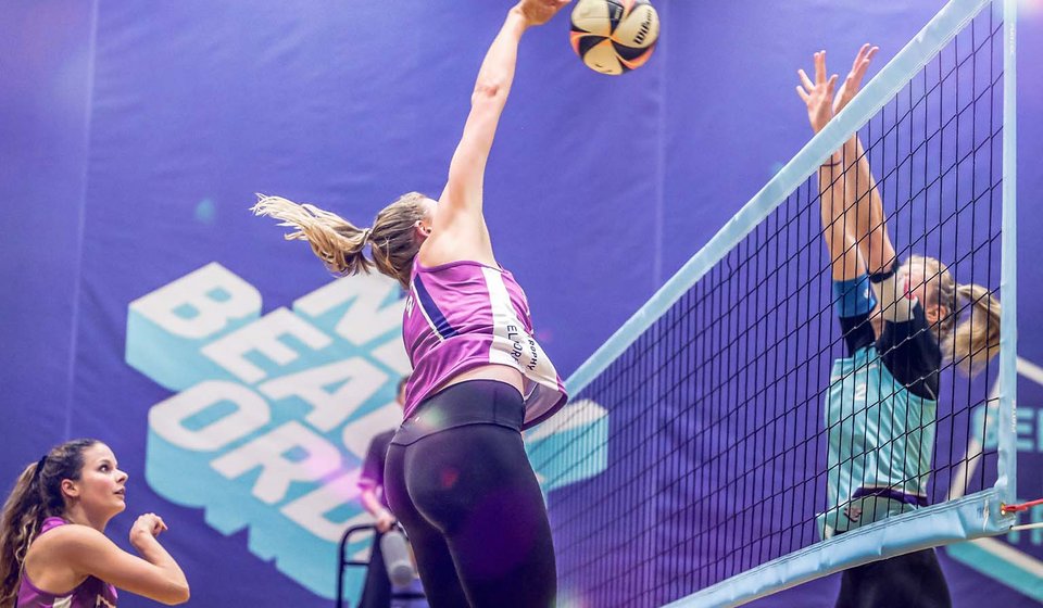 Eine Volleyball-Spielerin versucht den Ball über das Netz zu schlagen. Auf der anderen Seite vom Netz versucht eine andere Spielerin den Ball mit ausgestreckten Armen abzuwehren. 