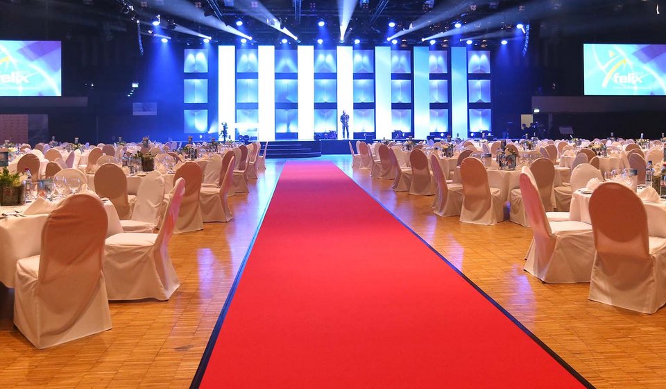 Blick aus dem Innenraum in Richtung der Bühne. In der Mitte liegt ein roter Teppich. Rechts und links stehen die unbesetzten Tische und Stühle. 