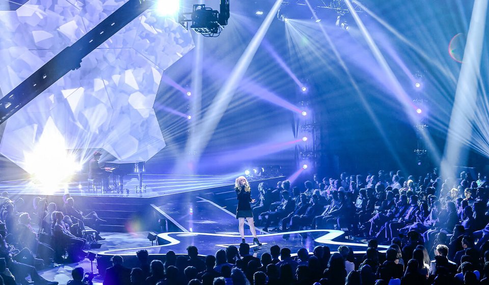 In der Mitte sieht man eine Sängerin auf einer sternenförmigen Bühne. Weiter hinten auf der Bühne sitzt ein Pianist. Um die Bühne herum sitzen Zuschauer:innen. Der Raum ist blau beleuchtet. 
