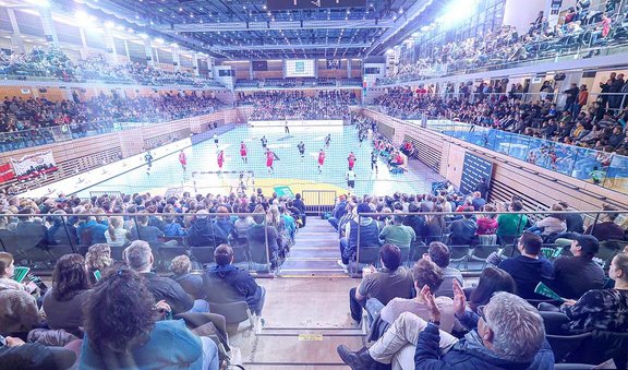 Blick von der Tribüne auf den Innenraum vom CASTELLO Düsseldorf. Im Innenraum ist ein Handballfeld aufgebaut, auf dem gerade ein Spiel läuft. Die Halle ist gefüllt mit Zuschauer:innen. 