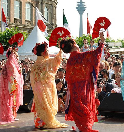 Das Bild zeigt drei Tänzerinnen in traditionellen japanischen Kimonos, die auf einer Bühne vor einem Publikum auftreten. Sie halten Fächer in ihren Händen und scheinen eine traditionelle japanische Tanzbewegung auszuführen. Die Kimonos sind farbenfroh, mit Mustern in Rot, Orange und Blau. Im Hintergrund sind Zuschauer zu sehen, die aufmerksam das Geschehen verfolgen. Im Hintergrund ragt der Rheinturm in Düsseldorf empor, was darauf hindeutet, dass diese Veranstaltung möglicherweise im Rahmen des Japan-Tages in Düsseldorf stattfindet, einem jährlichen Fest, das die deutsch-japanische Freundschaft feiert.