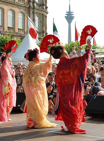 Das Bild zeigt drei Tänzerinnen in traditionellen japanischen Kimonos, die auf einer Bühne vor einem Publikum auftreten. Sie halten Fächer in ihren Händen und scheinen eine traditionelle japanische Tanzbewegung auszuführen. Die Kimonos sind farbenfroh, mit Mustern in Rot, Orange und Blau. Im Hintergrund sind Zuschauer zu sehen, die aufmerksam das Geschehen verfolgen. Im Hintergrund ragt der Rheinturm in Düsseldorf empor, was darauf hindeutet, dass diese Veranstaltung möglicherweise im Rahmen des Japan-Tages in Düsseldorf stattfindet, einem jährlichen Fest, das die deutsch-japanische Freundschaft feiert.
