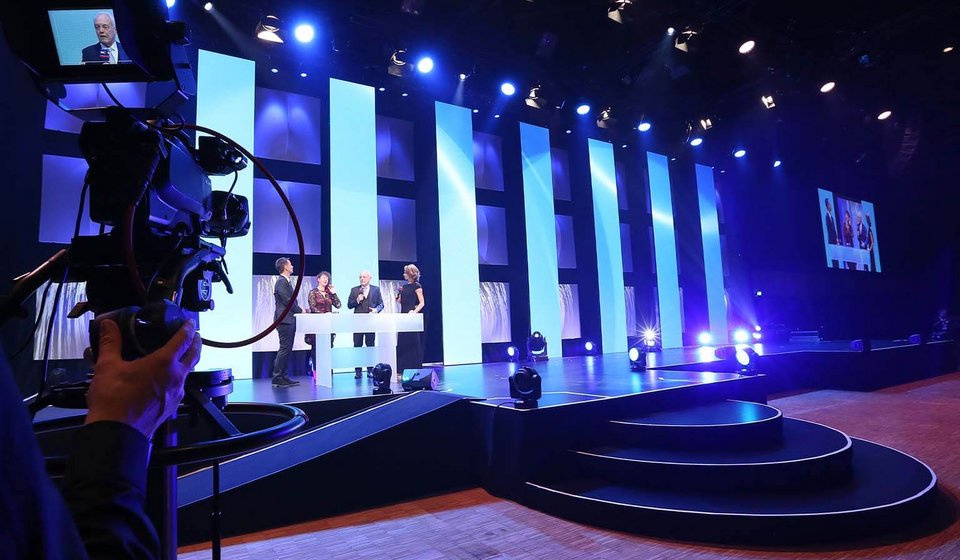 Blick auf die Bühne, auf der vier Personen an einem Moderationstisch stehen. Links sieht man eine Person an einer Filmkamera, die das Ganze aufnimmt. 