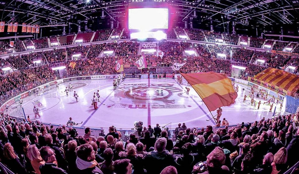 Blick auf das DEG Eishockeyfeld, auf dem sich die Spieler befinden. Am oberen Rand leuchtet der Videowürfel über der Eishockeyfläche. Rechts wird eine Fahne in den DEG Farben Rot und Geld geschwenkt. 
