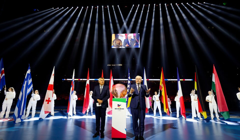 Das Bild zeigt eine bedeutende Eröffnungszeremonie der EHF EURO 2024 in der Merkur Spiel-Arena. Im Vordergrund stehen Michael Wiederer, der Präsident der Europäischen Handballföderation, der aufmerksam zuhört, und Frank-Walter Steinmeier, der Bundespräsident Deutschlands, der eine Rede hält. Sie sind umgeben von Flaggenträgern, die die Fahnen verschiedener europäischer Länder präsentieren. Über ihnen ist ein Bildschirm, der Steinmeier zeigt, und darüber strahlen helle Lichtkegel in den dunklen Raum und unterstreichen die feierliche Stimmung der Veranstaltung.
