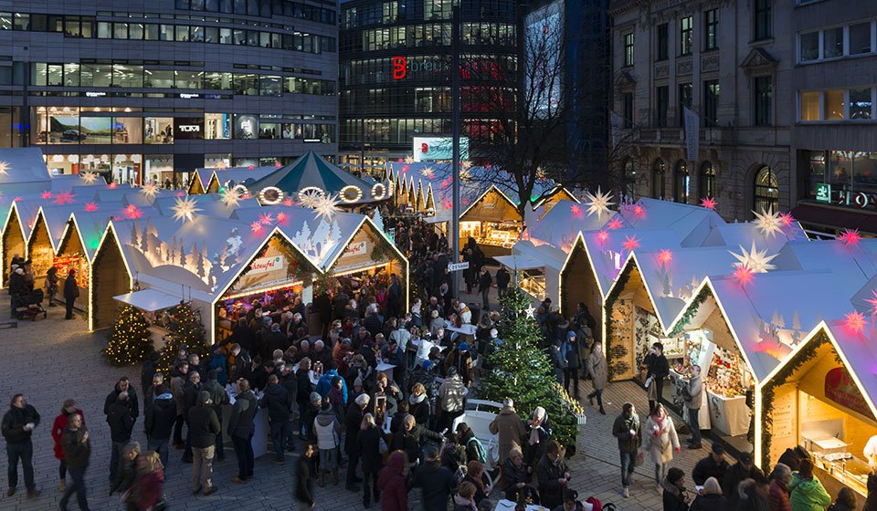 Weihnachtsmarkt in der Düsseldorfer Innenstadt: festliche Stände auf dem Schadowplatz bei weihnachtlicher Atmosphäre. Im Hintergrund Kö-Bogen mit Geschäften.