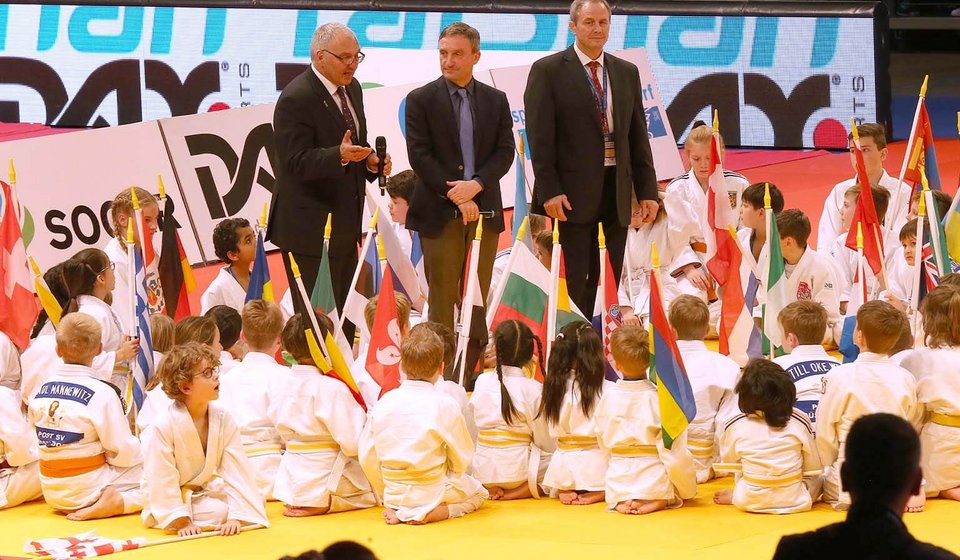 Kinder in Judo-Uniform und mit Fahnen in der Hand sitzen auf einer Matte im Kreis. In diesem Kreis stehen drei Männer in Anzügen und mit Mikrofonen. 