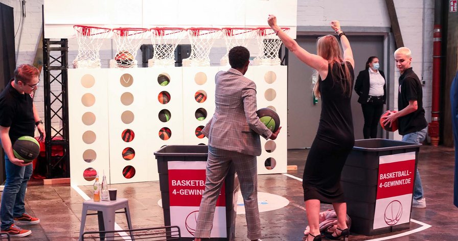 Vier Personen stehen vor einer Basketball-4-Gewinnt Wand. Eine Frau im langen Kleid jubelt, während sich ein Mann im Anzug für den Wurf bereitmacht. 