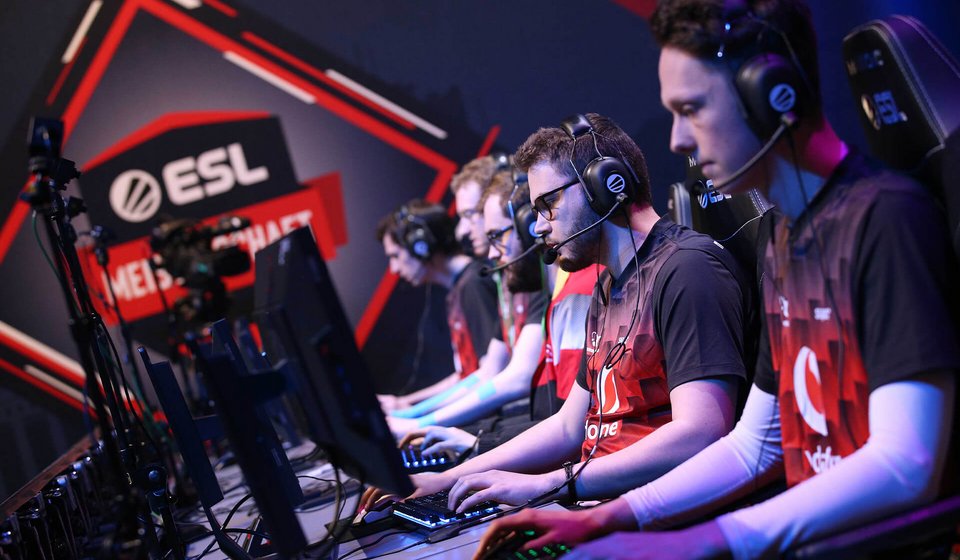 Spieler sitzen in einer Reihe vor Computern und haben Kopfhörer mit Mikrofon auf. Im Hintergrund sieht man das Logo der ESL Meisterschaft. 