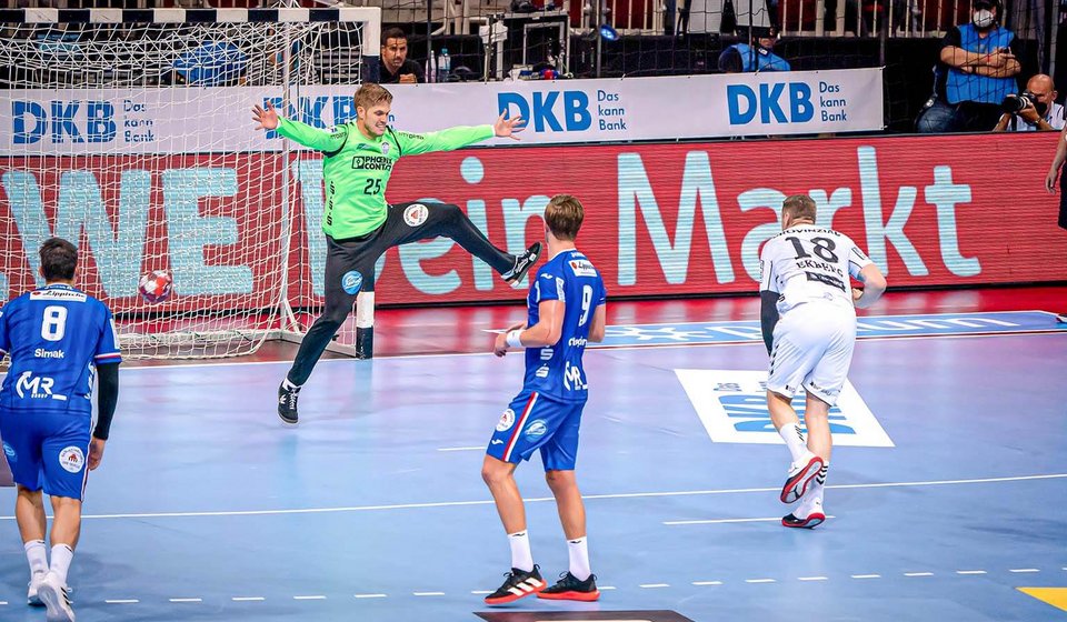 Man sieht vier Spieler auf einem Handballfeld. Ein Spieler ist gerade dabei den Ball auf das Tor zu werfen, wo der Torwart bereits springt um den Ball abzuwehren. 