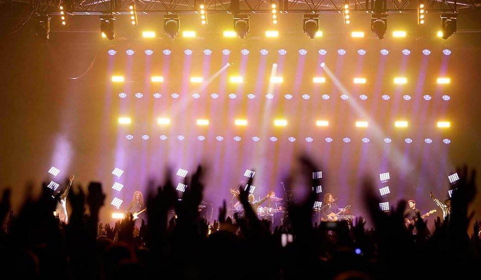 Blick aus der Menge auf die Bühne mit Lichtshow. Am unteren Rand sieht man verschwommen Hände, die in die Luft gestreckt sind.
