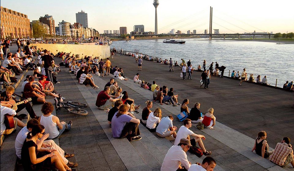 Im Vordergrund sieht man Menschen auf der Rheintreppe sitzen. Dahinter sieht man den Rhein und die Skyline von Düsseldorf mit dem Rheinturm und dem Medienhafen. 